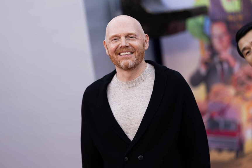 Bill Burr smiles in a grey shirt and black cardigan.