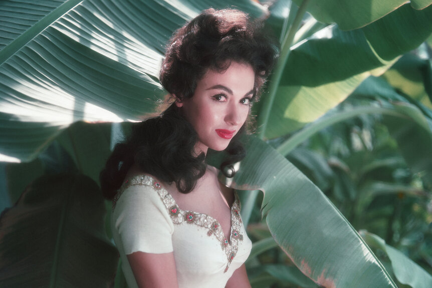 Rita Moreno stands in front of a large green plant in 1954