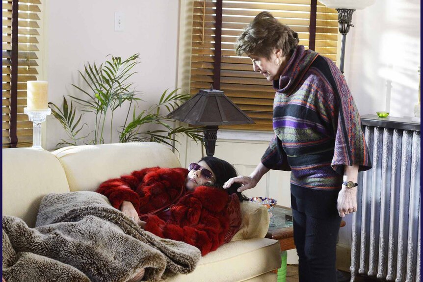 An older woman approaches Cecily Strong who is bundled up and laying on a couch.