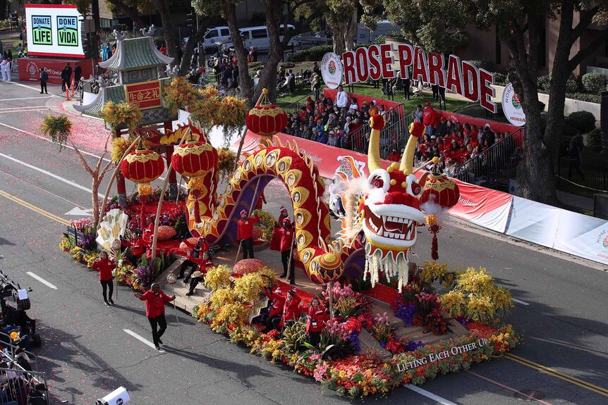 A floral arrangement parade float in the shape of a dragon is walked on a road.