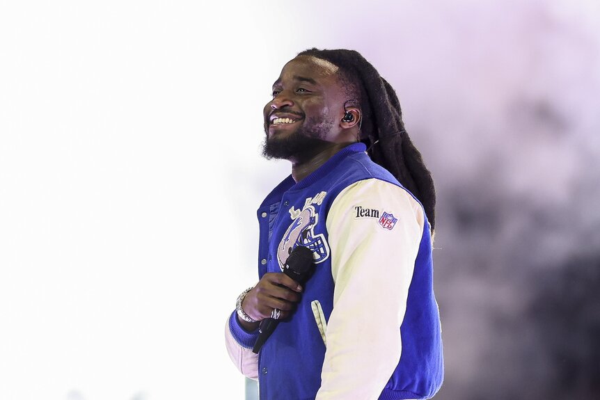 Shaboozey smiles in a blue and white NFL letterman jacket.