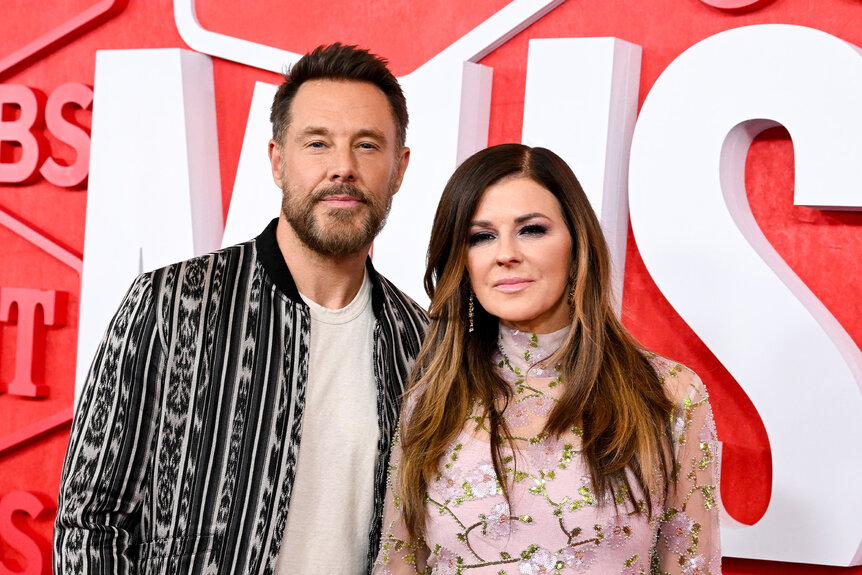 Jimi Westbrook And Karen Fairchild pose together in front of a red and white backdrop.