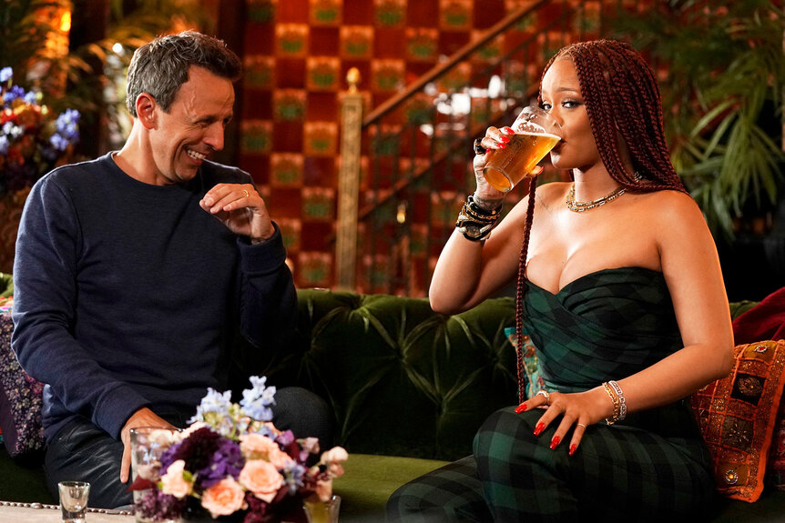 Seth Meyers and Rihanna drinking and laughing on a sofa.