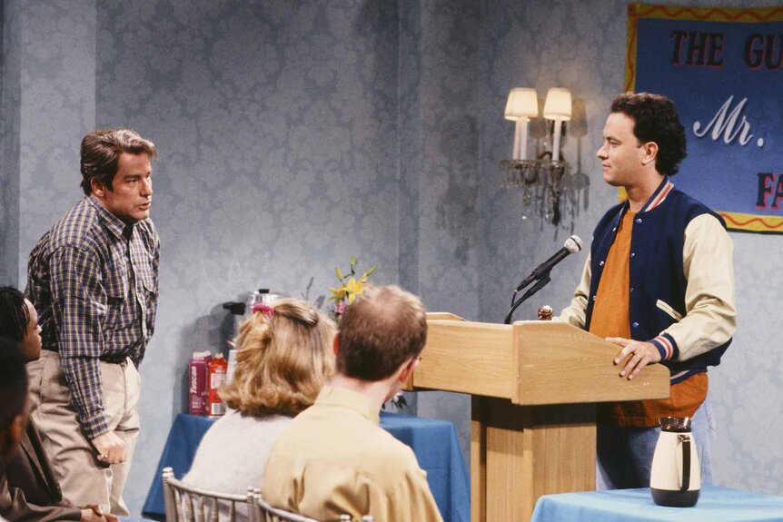 Phil Hartman stands to speak to Tom Hanks who stands at a podium on SNL.