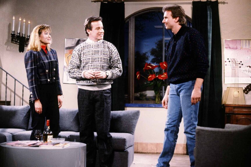 Leslie (Jan Hooks), Dan (Phil Hartman), and Stu (Joe Montana) stand in a living room during an SNL sketch.