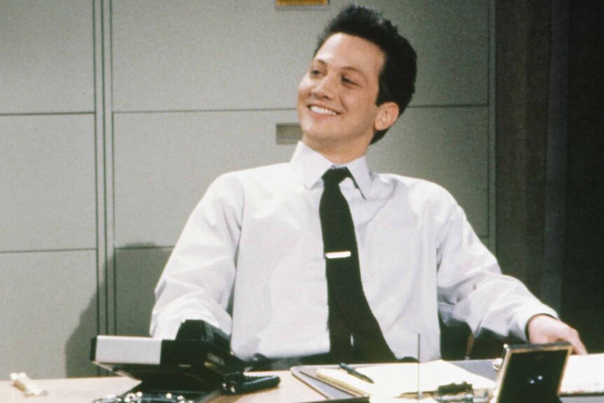 Richard Laymer (Rob Schneider) smiles behind a desk in a button down and tie during an SNL sketch.