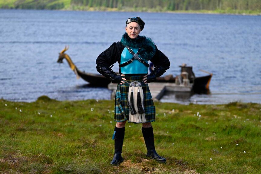 Alan Cumming wears a teal beret, feather top, and plaid blue skirt on The Traitors Season 3, Episode 1.