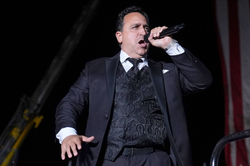 Christopher Macchio sings in a black suit on stage during a trump campaign rally