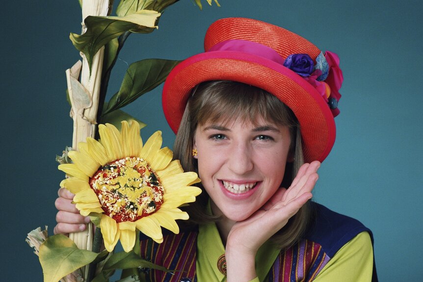 Blossom Russo (Mayim Bialik) wears an orange hat and smiles next to a sunflower Ion Blossom Season 1.