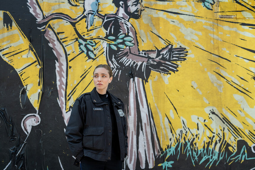 Mickey stands in front of a mural on a wall in Long Bright River Season 1, Episode 2.