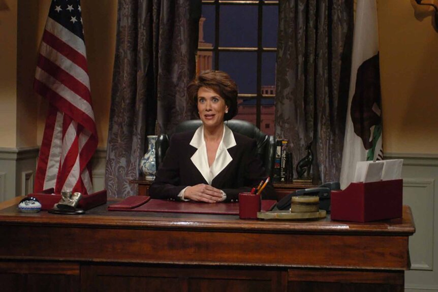 Nancy Pelosi (Kristen Wiig) sits at a desk flanked by a US flag and CA flag.