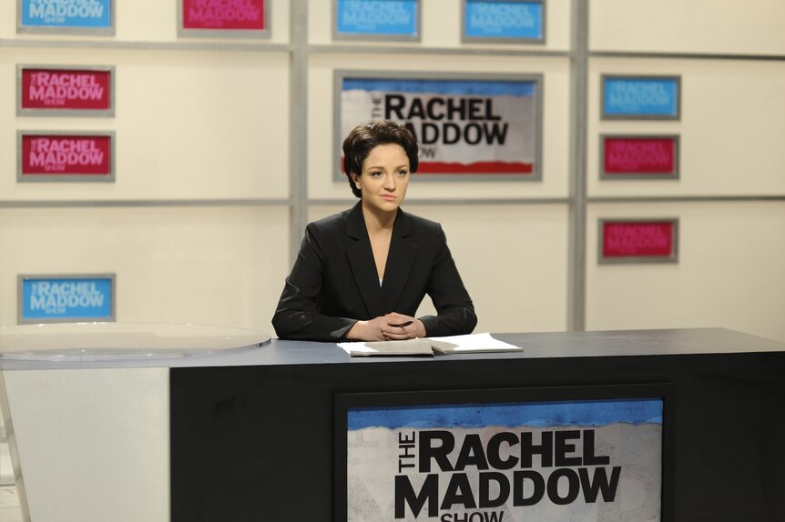 Rachel Maddow (Abby Elliott) sits at a newcast desk on SNL Episode 3412.