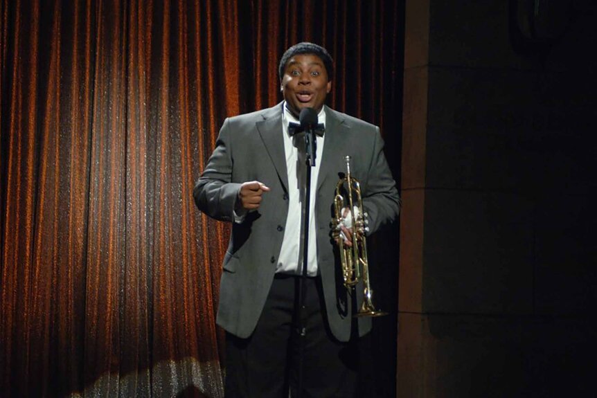 Louis Armstrong (Kenan Thompson) holds a trumpet and speaks into a mic on SNL Episode 3206.