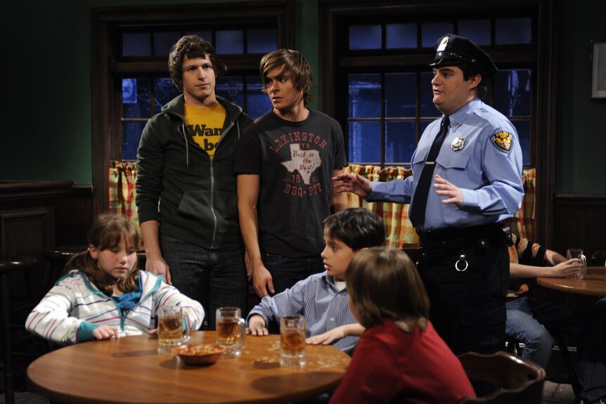 Patrons (Andy Samberg, Zac Efron) and Officer Williams (Bobby Moynihan) speak to a table of children during SNL Episode 3420.