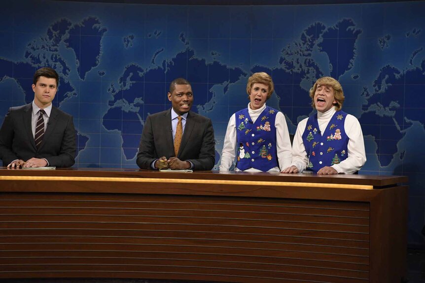 Colin Jost, Michael Che, Kat (Kristen Wiig), and Garth (Fred Armisen) sit at the Weekend Update desk on SNL Episode 1672.