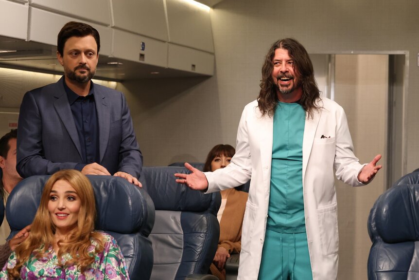 Chloe Fineman sits as Nate Bargatze and Dave Grohl walk around on a plane on Saturday Night Live Episode 1847.