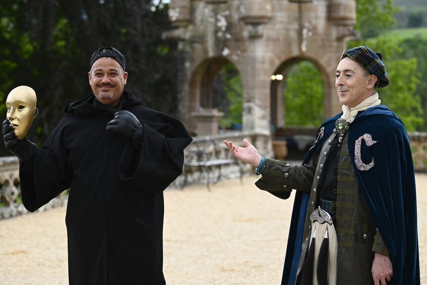 Alan Cumming gestures to Rob Mariano holding a mask while wearing a cloak on The Traitors Episode 303.