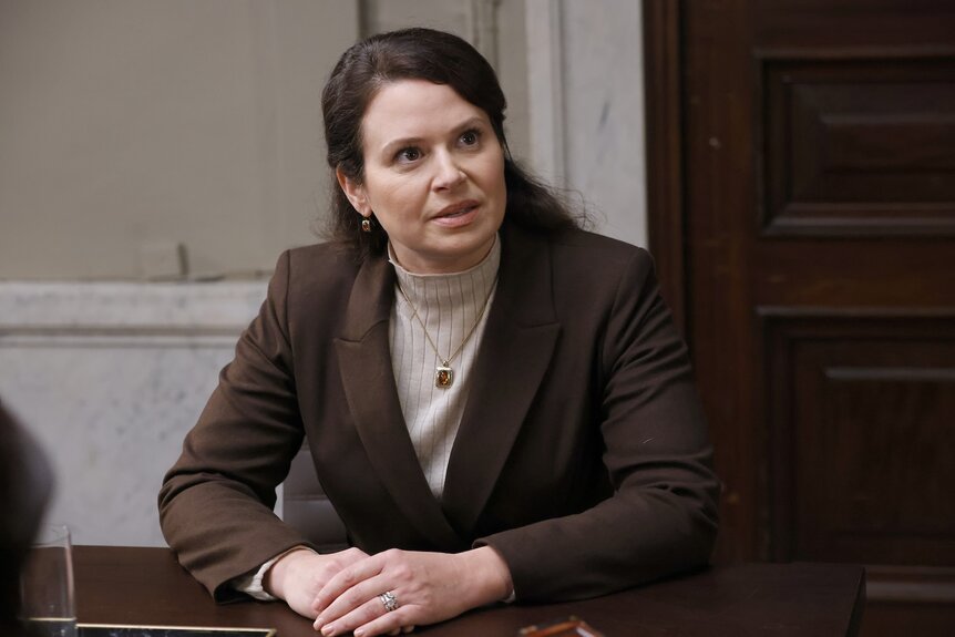 Victoria Beyer (Katie Lowes) folds her hands while wearing a brown suit.