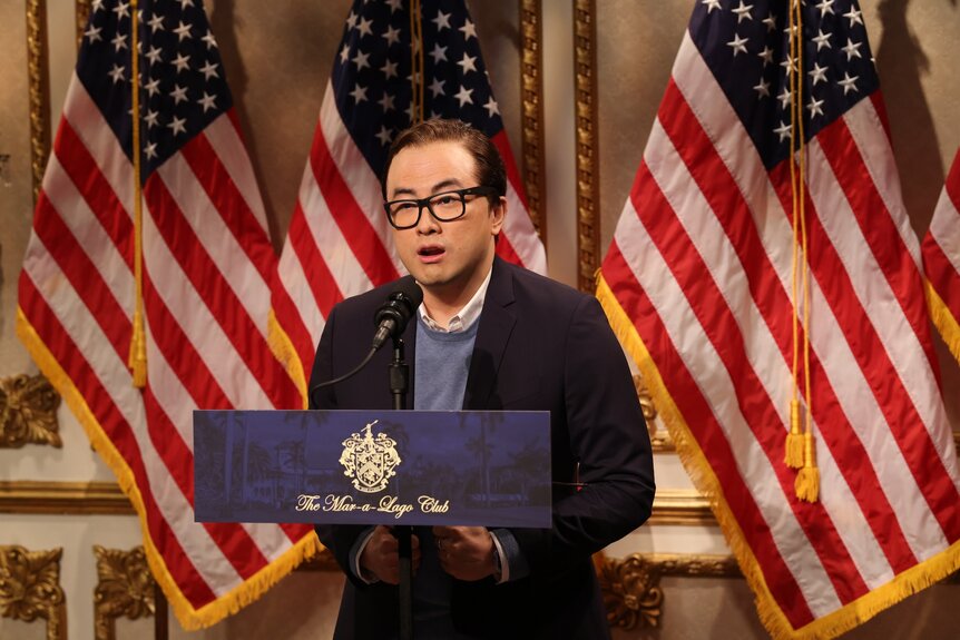 Bowen Yang in glasses standing behind a podium.