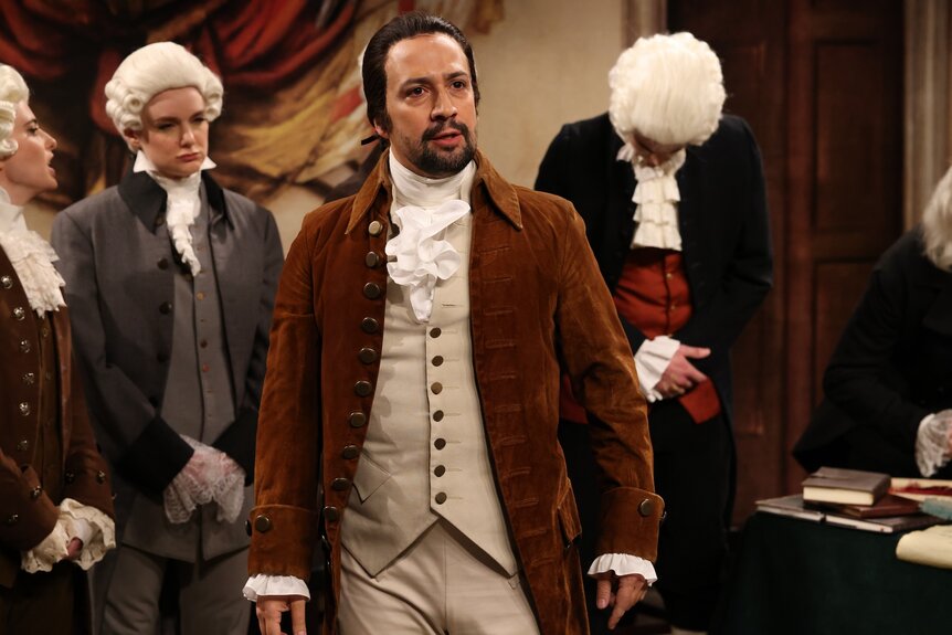 Lin-Manuel Miranda dressed as Alexander Hamilton next to people in white wigs.