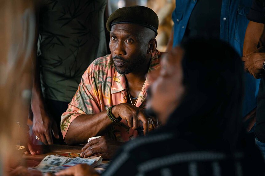 Duncan Kincaid (Mahershala Ali) sits with stacks of cash while people are around him in Jurassic World Rebirth (2025).