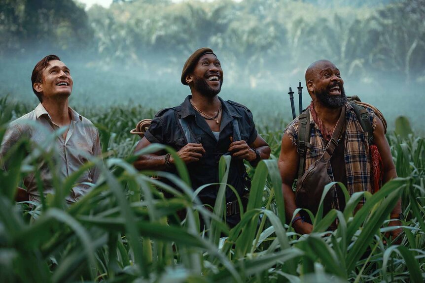 L to R: as Martin Krebs (Rupert Friend), Duncan Kincaid (Mahershala Ali), and Leclerc (Bechir Sylvain) smile and look up while standing in tall grass in Jurassic World Rebirth (2025).