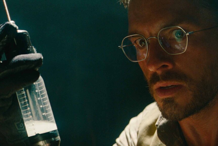 Dr. Henry Loomis (Jonathan Bailey) looks at a giant syringe full of white liquid in Jurassic World Rebirth (2025).