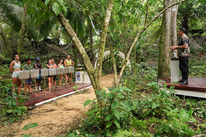 The cast stands next to briefcases in the forest on Deal or No Deal Island appears on Episode 208.