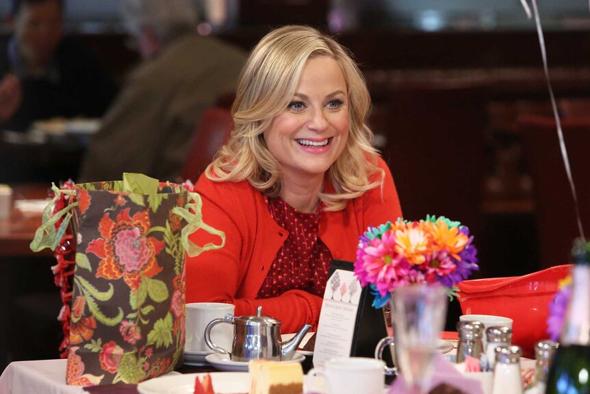 Leslie Knope sitting at a table with food and decorations on Parks and Recreation Season 6, Episode 17.