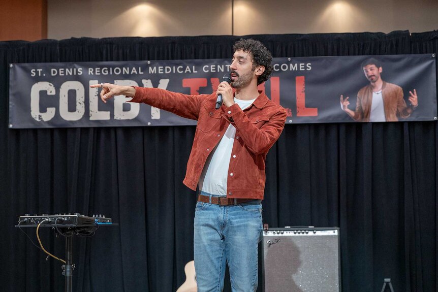 Colby Twill on stage while talking on a microphone in St. Denis Medical Season 1, Episode 13.