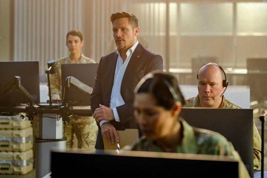 Oliver Odell standing among a group of people at their desks in The Hunting Party Season 1, Episode 2.