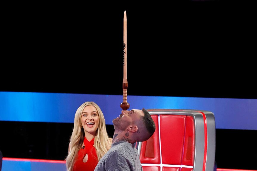 Adam Levine balancing a sword on his head while Kelsea Ballerini watches on The Voice Season 27, Episode 3.