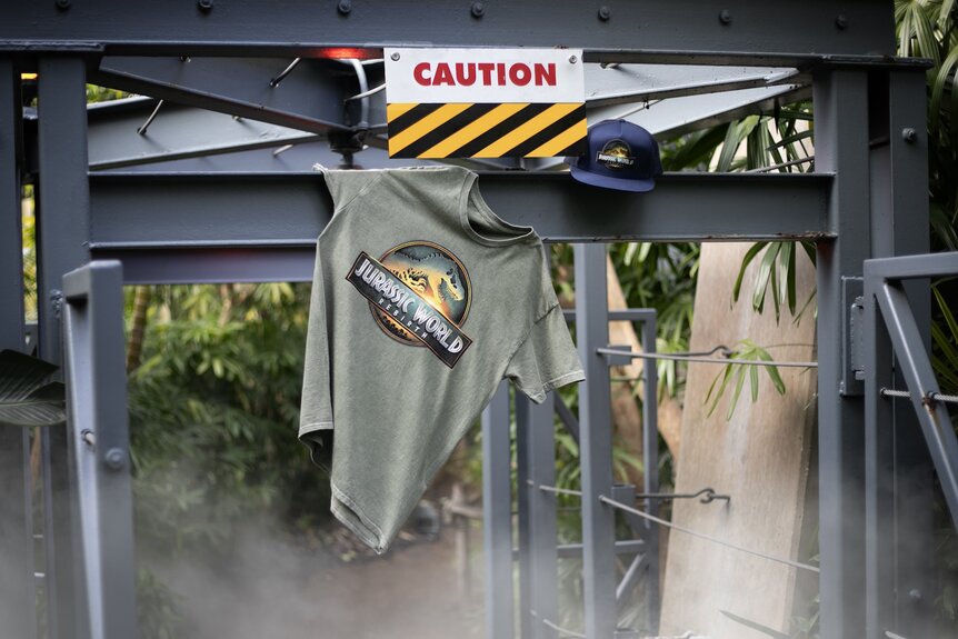 A Jurassic World: Rebirth (2025) shirt and hat hang near a "Caution" sign.