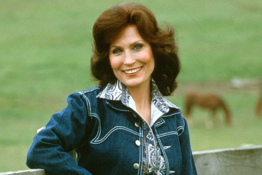 Loretta Lynn smiling outdoors and leaning on a fence.