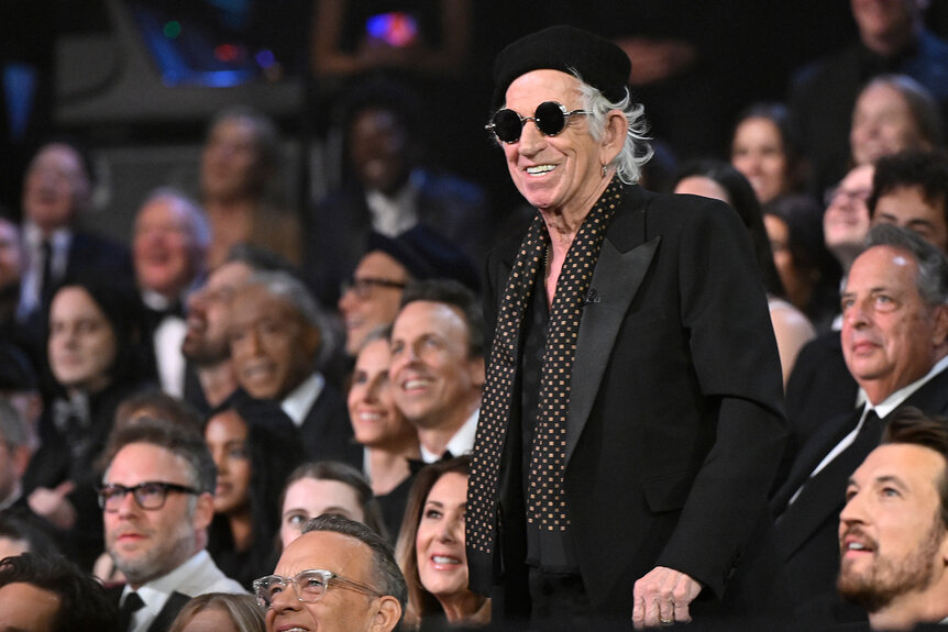 Keith Richards stands in a black outfit as Seth Rogan, Seth Meyers, and Miles Teller sit in the audience of the SNL50 special.