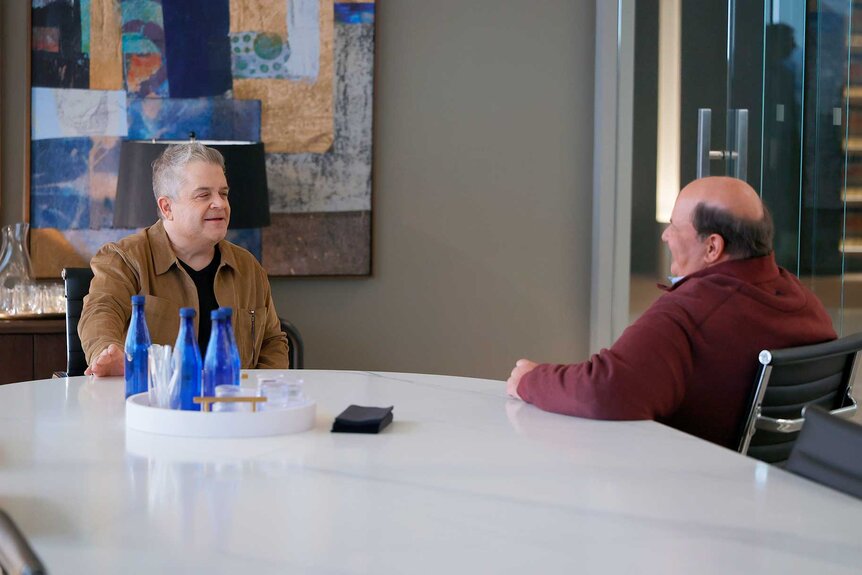 Patton Oswalt sitting at a desk talking to Brian Baumgartner on Suits LA Season 1, Episode 3.